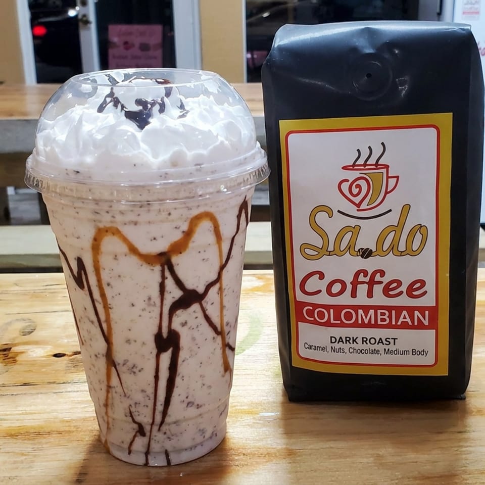 Chocolate-drizzled whipped cream coffee shake beside a bag of Sando Coffee Colombian dark roast on a wooden café table.