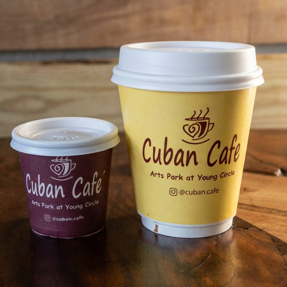 Two lidded Cuban Café takeaway cups, one purple and one yellow, sit on a rustic wooden table.