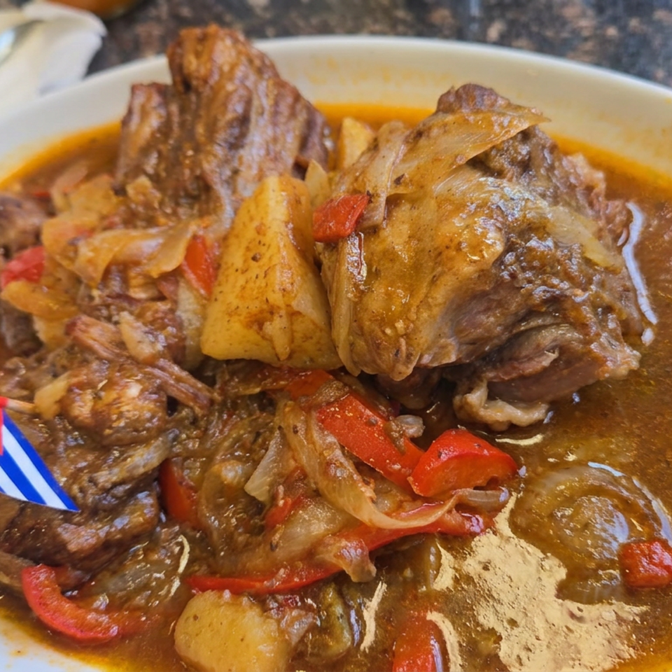 Stewed beef with potatoes, onions, and red peppers in glossy brown gravy, served on a white plate with a small Cuban flag pick.