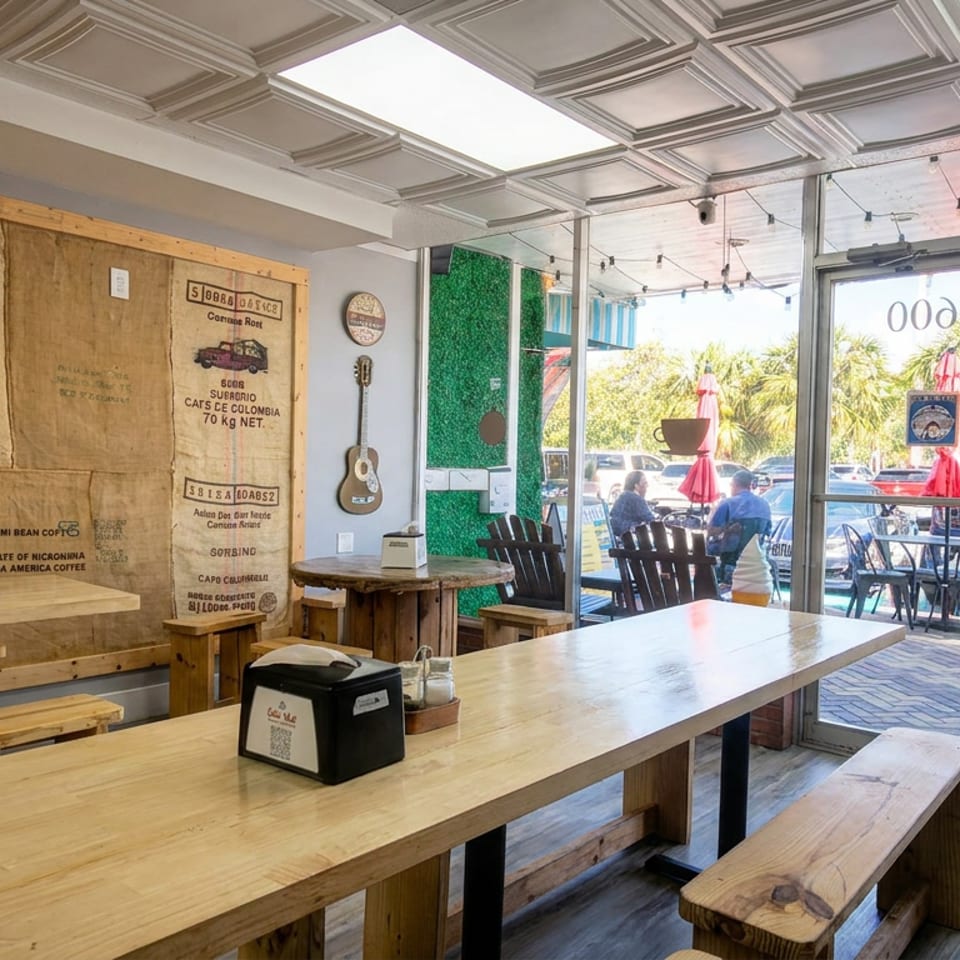 Bright rustic café interior with communal wooden tables, coffee sack wall decor, hanging guitar, and sunny outdoor patio seating.
