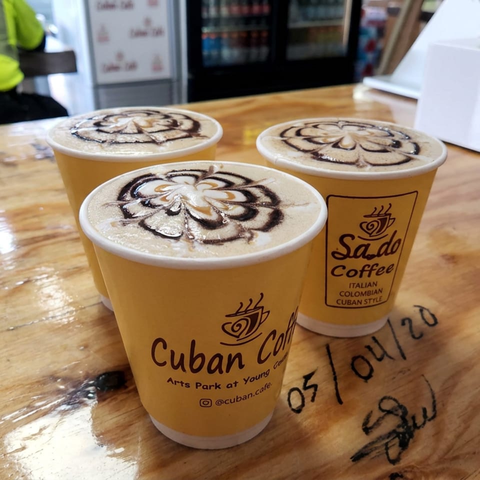 Three Cuban Coffee cups with decorative foam art sit on a wooden café counter.