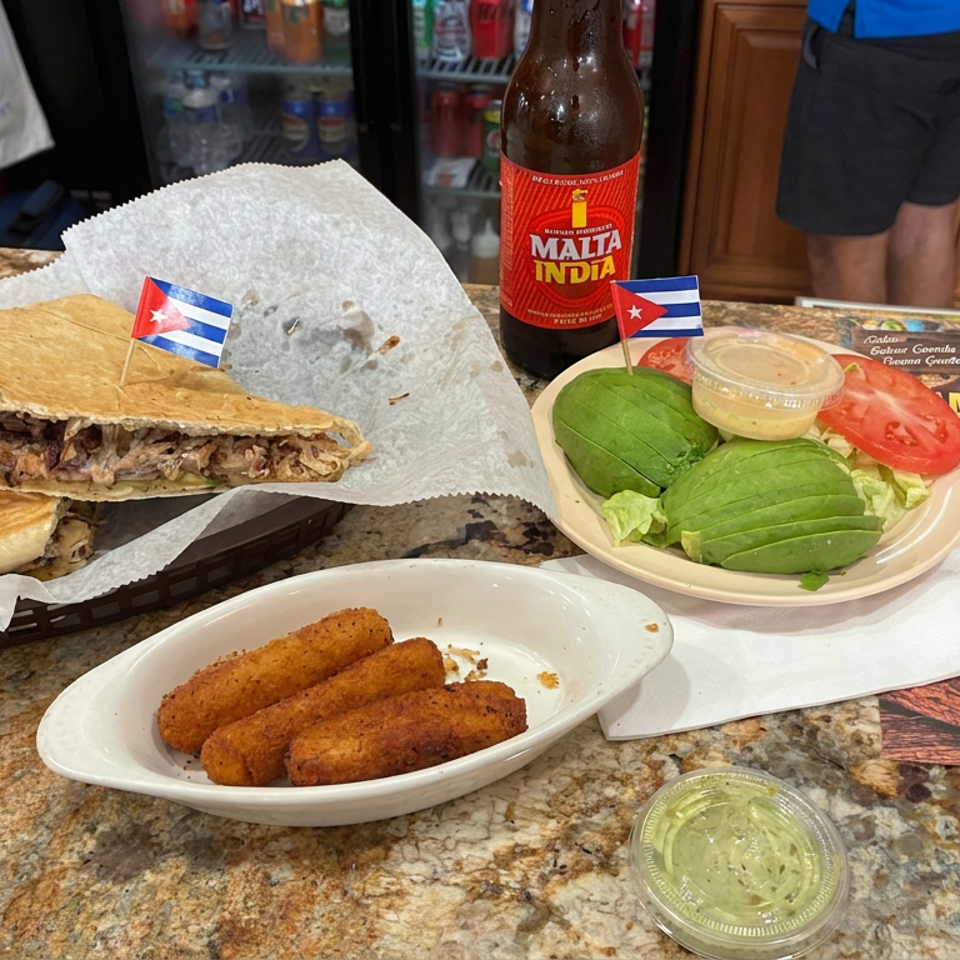 Cuban meal featuring a pressed sandwich, croquettes, avocado salad, Malta India soda, and Cuban flags on a restaurant counter.